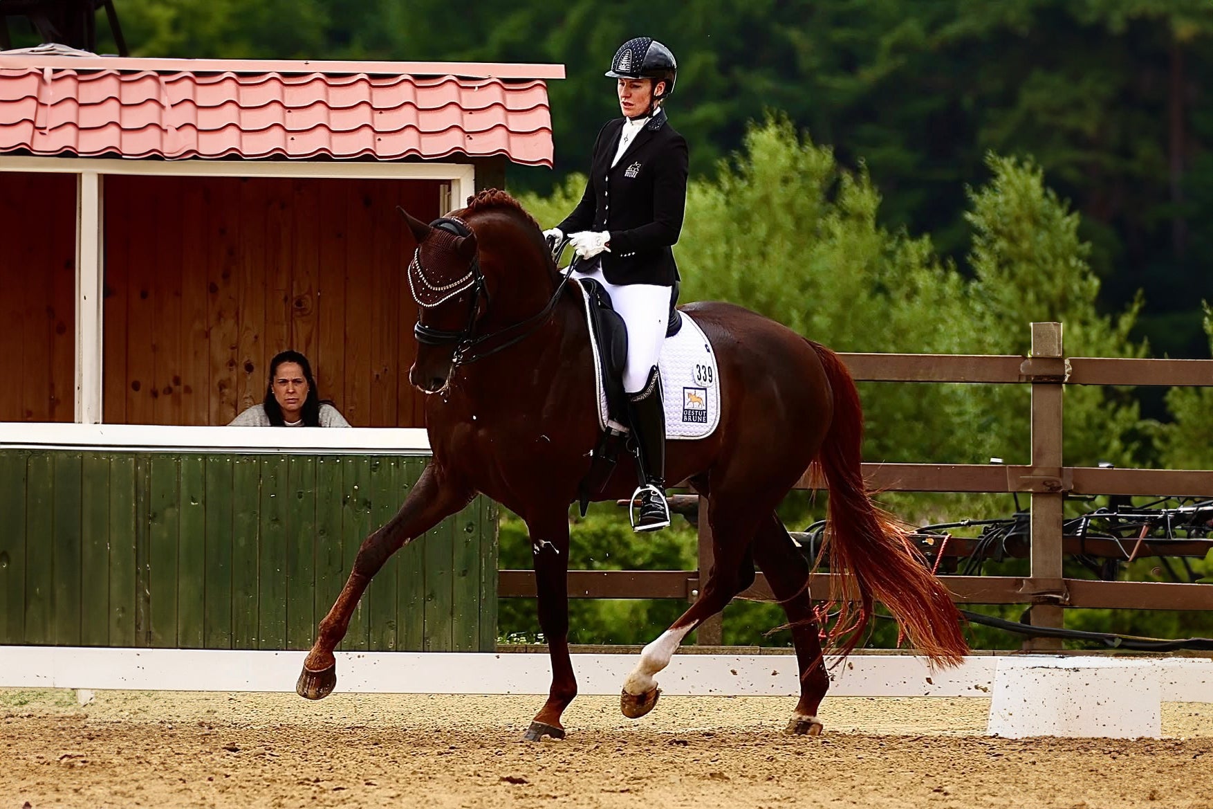 Bernadette Brune trotting her horse in a dressage test at an outdoor show, wearing a JuulC competition jacket